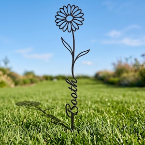 May include: A black metal garden stake featuring a stylized daisy flower at the top and the name Booth written in a cursive script along the stem. The decorative piece is designed to be inserted into the ground for outdoor display.