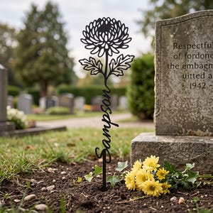 May include: A black metal garden stake featuring a cutout design of a chrysanthemum flower at the top and the name Sampson written in a cursive script along the stem, positioned vertically in the soil near a stone grave marker.