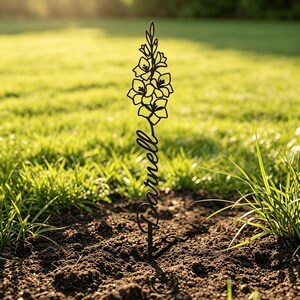 May include: A black metal garden stake featuring a cutout design of a tall flower stalk at the top and the name Darnell written in a cursive script font below the floral element, with a single pointed stake base inserted into the dark soil.
