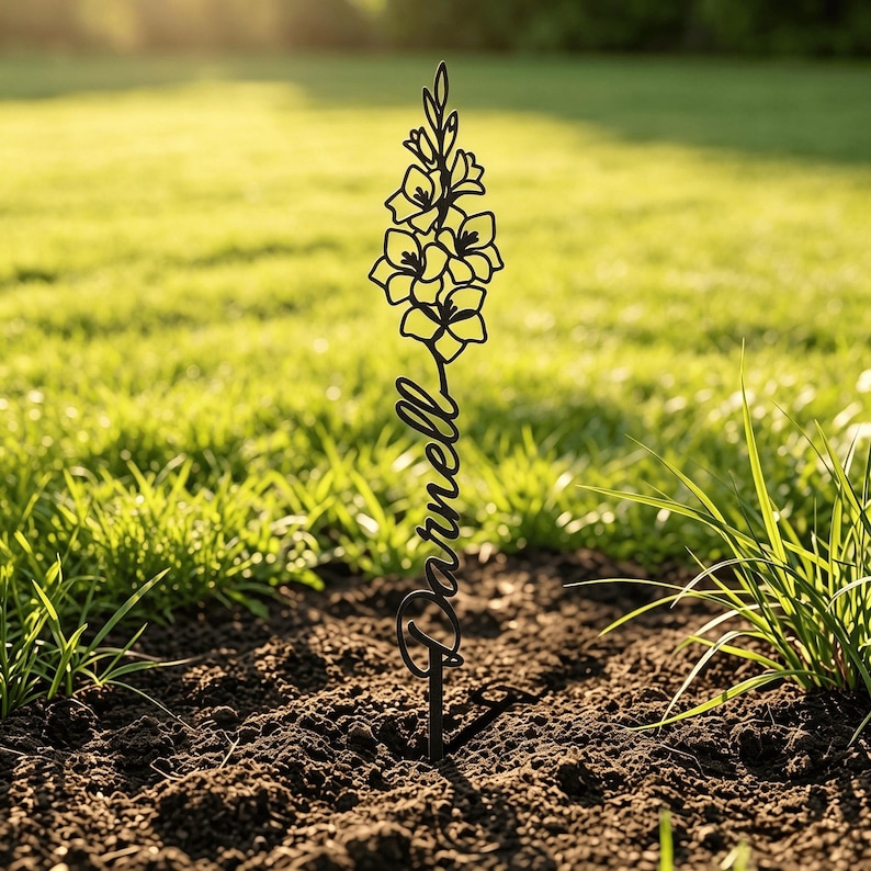 May include: A black metal garden stake featuring a cutout design of a tall flower stalk at the top and the name Darnell written in a cursive script font below the floral element, with a single pointed stake base inserted into the dark soil.