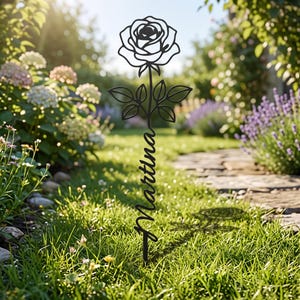 May include: A black metal garden stake featuring a stylized rose flower at the top and the name Martina written in a cursive script along the stem. The decorative piece is designed to be inserted into the ground as a personalized garden marker.