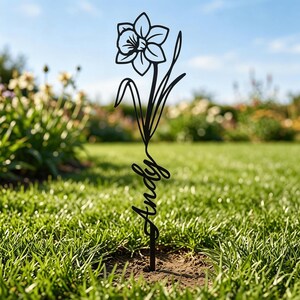 May include: A black metal garden stake featuring a cutout design of a single daffodil flower at the top and the name Andy written in a cursive script font along the stem, positioned vertically in the ground.