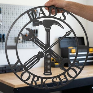 Può includere: Un cartello in metallo nero con le parole "DADS WORKSHOP" in un design circolare. Il cartello presenta un martello, una chiave inglese, un cacciavite e un lesina, tutti in una disposizione incrociata, che simboleggia un tema di officina.