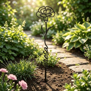 May include: A black metal garden stake featuring a stylized carnation flower at the top and the name Teresa written in a cursive script along the stem. The stake is inserted into the soil, standing upright among various green garden plants.