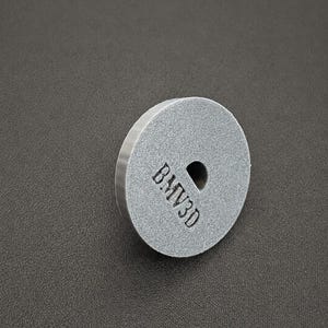 May include: A circular, gray, textured disc with a flat face and a beveled edge. The front surface features a small, centered circular hole and the embossed text BMV3D positioned vertically to the left of the center hole.