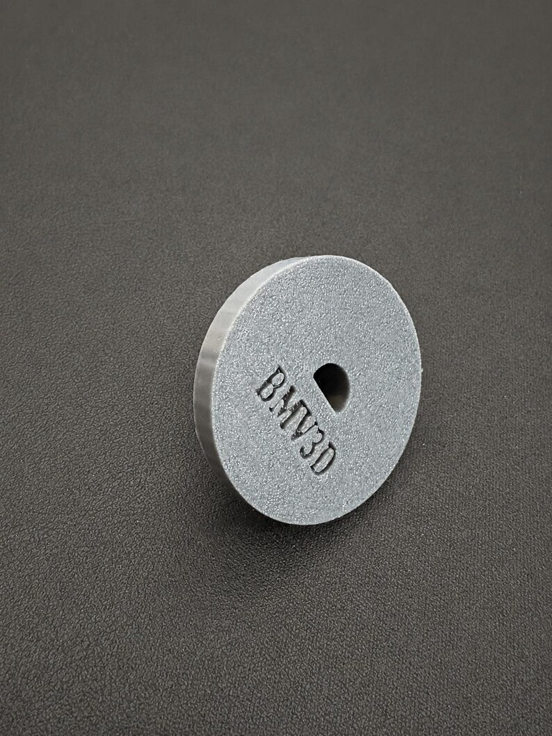 May include: A circular, gray, textured disc with a flat face and a beveled edge. The front surface features a small, centered circular hole and the embossed text BMV3D positioned vertically to the left of the center hole.