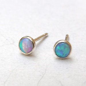 Pode incluir: Um par de brincos pequenos com pedras de opala iridescentes em prata. As opalas exibem uma mistura de tons azuis, verdes e rosa. Os brincos têm um design simples e minimalista.