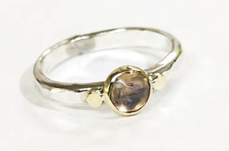 May include: A silver ring with a hammered finish and a round, brown gemstone set in a gold bezel. The ring has two smaller gold accents on either side of the gemstone.