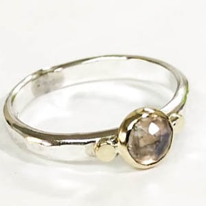 May include: A silver ring with a hammered finish and a round, clear gemstone set in gold. The ring has two small gold accents on either side of the gemstone.