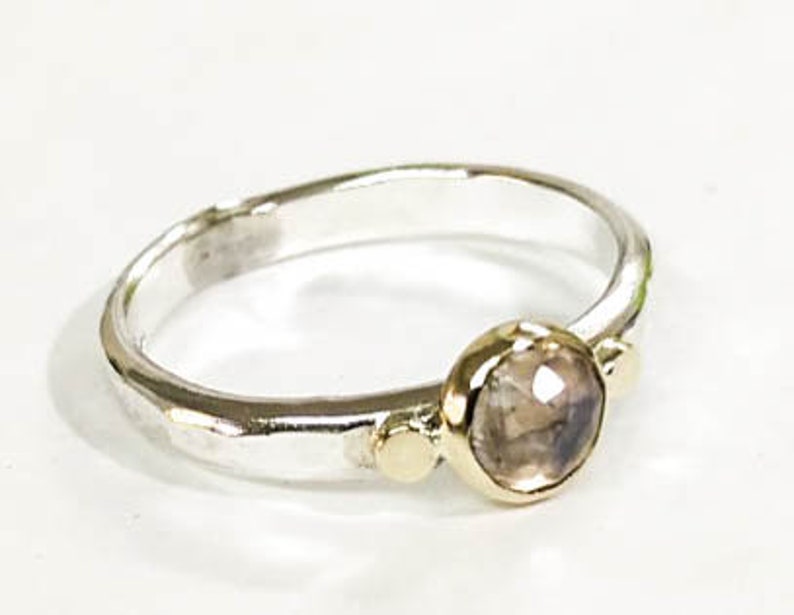 May include: A silver ring with a hammered finish and a round, clear gemstone set in gold. The ring has two small gold accents on either side of the gemstone.