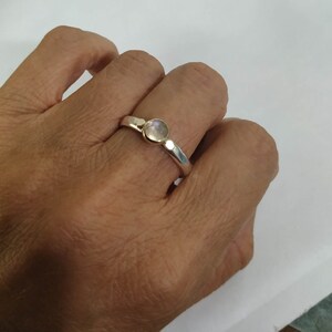 May include: A silver ring with a round, clear gemstone set in the center. The ring is worn on a person's finger.
