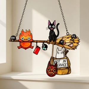 Könnte beinhalten: Ein hängendes Buntglas-Ornament, das einen Besenstiel mit einer schwarzen Katze, einem Feuergeist und kleinen Rußmännchen zeigt. Eine beige Tasche mit der Aufschrift NOT IN SERVICE hängt am Besen, ergänzt durch eine rote Maske und ein rotes Etikett.