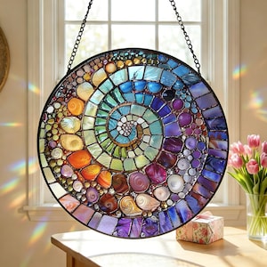 May include: A circular stained glass suncatcher featuring a vibrant spiral pattern of multicolored glass pieces in shades of blue, purple, orange, and yellow, encased in a dark metal frame and suspended by a black chain in front of a window.