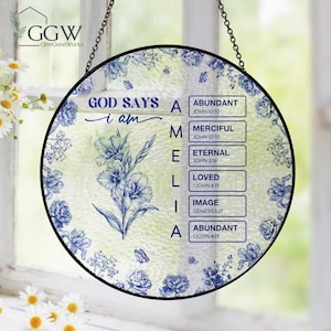Puede incluir: Vidriera redonda con marco negro. Presenta diseños florales azules y el texto "GOD SAYS I AM MELIA" con referencias bíblicas. Incluye una cadena para colgar, ideal para decoración del hogar.
