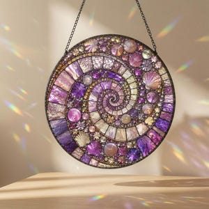 May include: A circular hanging suncatcher featuring a spiral mosaic design composed of various purple, lavender, and iridescent glass pieces, including small shell-like accents and round beads, suspended by a thin black metal chain.