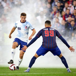 May include: Two soccer players in action on a green field. One player wears a white and light blue jersey with the number 99, blue shorts, and white socks. The other player has a navy blue jersey with red stripes, the number 99, and the text "YOUR NAME".