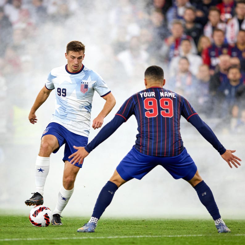 May include: Two soccer players in action on a green field. One player wears a white and light blue jersey with the number 99, blue shorts, and white socks. The other player has a navy blue jersey with red stripes, the number 99, and the text "YOUR NAME".