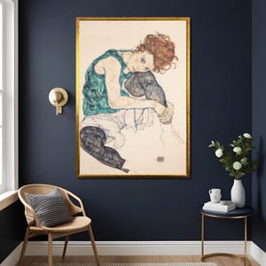 May include: A framed artwork featuring a seated figure in a contemplative pose. The figure is depicted with red hair, wearing a green top and dark pants. The artwork is in a gold frame, displayed against a dark blue wall, with a chair and side table in the room.