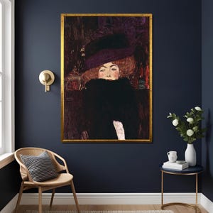 Può includere: Ritratto incorniciato di una donna con un grande cappello nero e una stola di pelliccia nera. Il dipinto, in una cornice dorata, è appeso a una parete blu scuro. La donna ha i capelli rossi e un incarnato pallido.