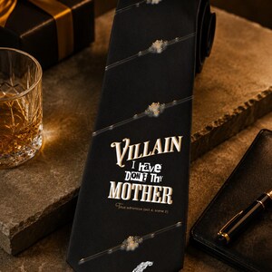 Op de afbeelding: Zwarte stropdas met de witte tekst "Villain I Have Don't Thy Mother", gouden accenten en een veerillustratie. De stropdas heeft diagonale gouden strepen. Waarschijnlijk gemaakt van zijde of een vergelijkbaar materiaal.