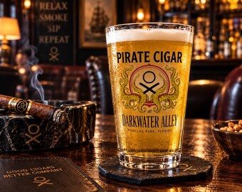May include: A clear glass pint filled with golden beer, featuring the text PIRATE CIGAR at DARKWATER ALLEY, PINELLAS PARK, FLORIDA, and a logo with green tentacles. A cigar rests on a black marble ashtray nearby, with a black coaster underneath.