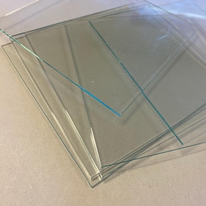 May include: A stack of multiple clear, rectangular glass sheets with smooth, straight edges. The transparent panes are layered at slight angles, revealing their thin profile and reflective surfaces. Each individual sheet is colorless and flat.