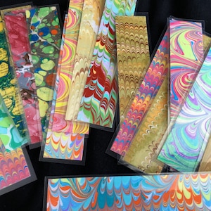 May include: A collection of colourful marbled paper bookmarks with various patterns and designs. The bookmarks are in a variety of colours, including red, green, blue, yellow, and orange. The bookmarks are all rectangular in shape and are approximately 25 cm long.