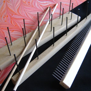 May include: Two wooden boards with rows of metal pins, used for marbling paper. A long wooden dowel with a white handle is resting on the pins. A smaller wooden board with a row of metal pins is also shown.