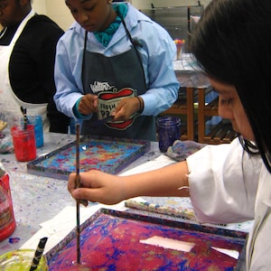 May include: Two people are working on a marbling art project. One person is using a stick to swirl the paint in a tray. The tray is filled with a pink, blue, and white marbled design. The tray is on a table with a white surface. The table is covered in paint splatters. The person wearing a white lab coat has a blue and white striped apron with the words "Walters Fresh Produce" printed on it.