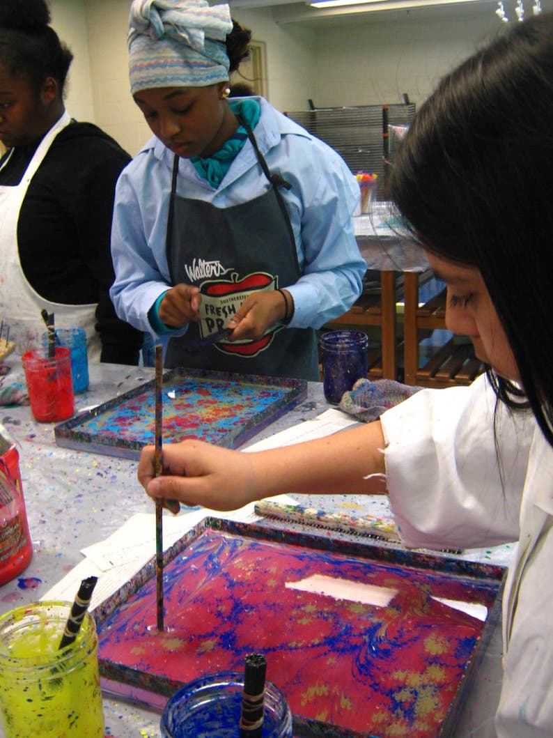 May include: Two people are working on a marbling art project. One person is using a stick to swirl the paint in a tray. The tray is filled with a pink, blue, and white marbled design. The tray is on a table with a white surface. The table is covered in paint splatters. The person wearing a white lab coat has a blue and white striped apron with the words "Walters Fresh Produce" printed on it.