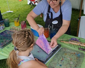 May include: A young girl wearing a white apron with colorful paint splatters watches as an adult woman demonstrates marbling paper. The woman is holding a piece of paper with pink, purple, and blue marbled patterns over a tray with a clear plastic surface. The woman is wearing a black apron with the word "Marbling" printed on it. The sign behind the woman reads "marblingsun" and "Genuinely Handmade".