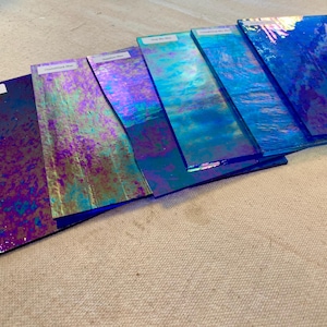 Könnte beinhalten: Eine Sammlung von sechs rechteckigen Glasscheiben, die fächerförmig angeordnet sind. Jede Scheibe weist eine ausgeprägte irisierende Oberfläche mit verschiedenen Blau-, Violett- und Türkistönen auf. Auf jeder Glasscheibe befindet sich ein kleines weißes Etikett.