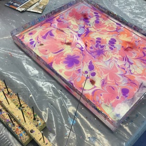 May include: A tray with a clear plastic top filled with water and swirling pink, purple, and yellow paint. A wooden tool with metal pins is on the left side of the tray.