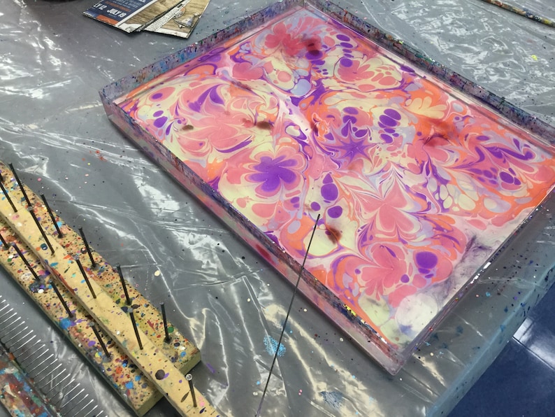 May include: A tray with a clear plastic top filled with water and swirling pink, purple, and yellow paint. A wooden tool with metal pins is on the left side of the tray.