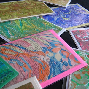 Puede incluir: Una colección de tarjetas de papel marmoleado de colores con una tarjeta rosa en la parte superior que presenta un extracto de texto sobre los suburbios. El texto está en azul y naranja con un fondo marmoleado.