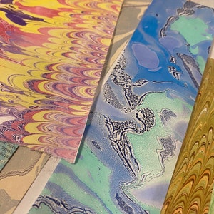 May include: Close-up of four different marbled paper samples. The papers are in shades of yellow, red, blue, green, and white. The marbling is intricate and detailed.