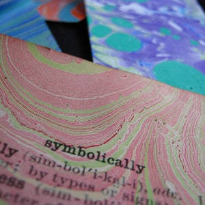May include: Close-up of a dictionary page with the word "symbolically" defined. The page has a pink and green marbled pattern.