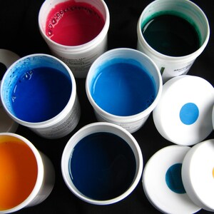 May include: Assortment of paint colors in small containers. The colors include red, blue, green, and yellow. The containers are white with black lids.