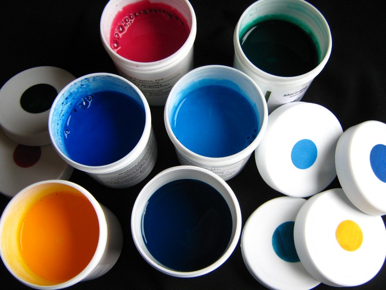 May include: Assortment of paint colors in small containers. The colors include red, blue, green, and yellow. The containers are white with black lids.