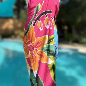 May include: A vibrant pink sarong with a large floral design in orange, yellow, and green. The sarong is draped over a mannequin, showcasing the colorful botanical print. The background features a swimming pool.