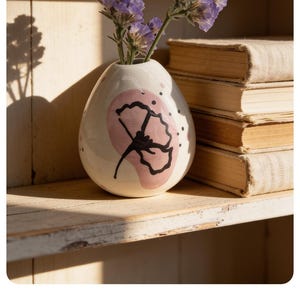 May include: A small, egg-shaped white ceramic vase featuring a hand-painted pink circular accent and a black line-art floral design. The vase contains a few sprigs of purple statice flowers and is positioned next to a stack of three old books.