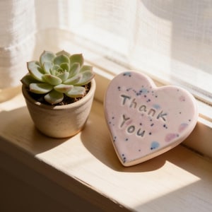 May include: A small, light-coloured ceramic pot containing a green succulent plant next to a heart-shaped, pale pink ceramic plaque with blue and pink speckles. The plaque features the embossed text Thank You in a simple, dark-coloured font.