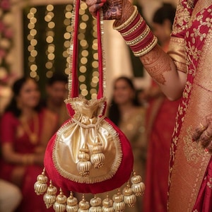 Puede incluir: Una bolsa roja y dorada con cordón y borlas doradas. La bolsa tiene forma redonda con una parte delantera de satén dorado y una parte trasera de terciopelo rojo. El asa es roja con ribete dorado.