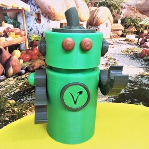 May include: A green robot figurine with brown eyes and a circular emblem with a V-shaped design. The robot has a cylindrical body, arms, and a head with a rounded top. The background features a colorful, whimsical scene.