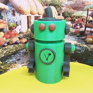 May include: A green robot toy with brown eyes and a matching hat. The robot has gray arms and a circular emblem with a stylized V on its chest. The toy is sitting on a yellow surface with a painted background of a market scene.