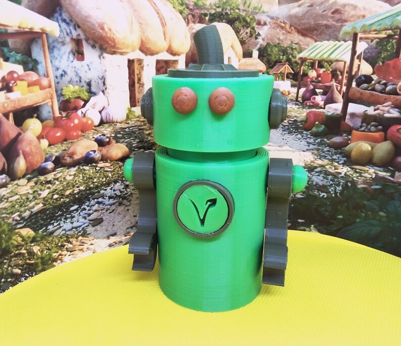 May include: A green robot toy with brown eyes and a matching hat. The robot has gray arms and a circular emblem with a stylized V on its chest. The toy is sitting on a yellow surface with a painted background of a market scene.