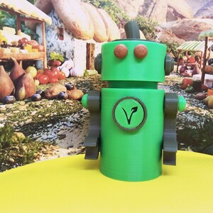 May include: A green robot figurine with brown accents, including arms, eyes, and a circular emblem on its chest. The emblem features a stylized leaf design. The robot is set against a backdrop of a market scene with food items.