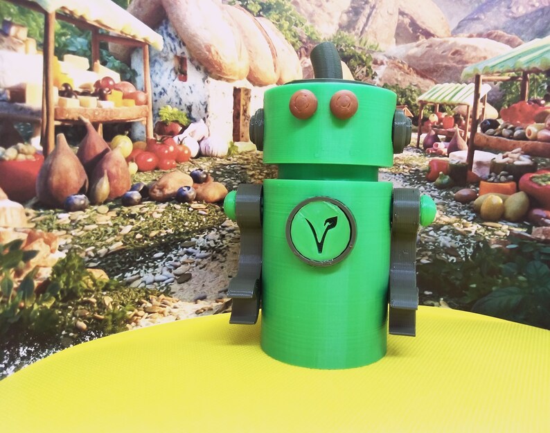 May include: A green robot figurine with brown accents, including arms, eyes, and a circular emblem on its chest. The emblem features a stylized leaf design. The robot is set against a backdrop of a market scene with food items.