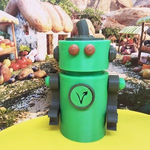May include: A green robot toy with brown arms, eyes, and a hat. The robot has a circular emblem on its chest with a stylized arrow. The background features a whimsical village scene with food stalls and a small house.