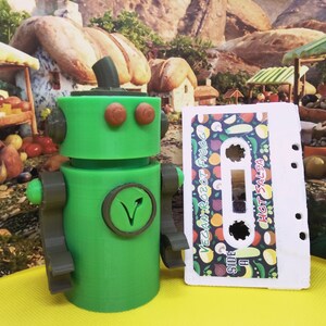 May include: A green robot figure with brown accents and a circular emblem. Beside it is a cassette tape labeled "Vegan Robot Food" and "Hot Salsa" with a colorful vegetable pattern. The robot has a plant-based symbol.
